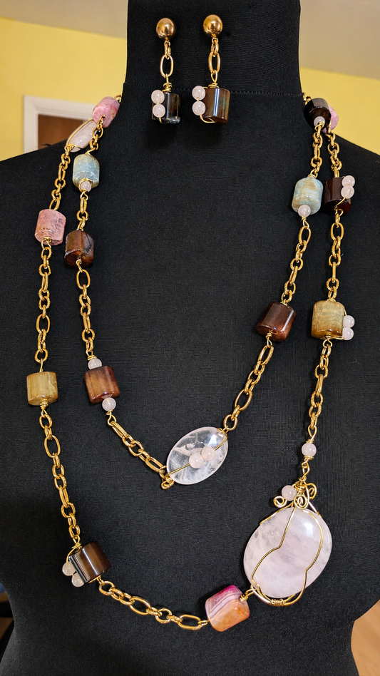 A gold-tone layered necklace and matching drop earrings displayed on a black mannequin bust. The necklace features a gold chain with multiple strands, mixed-color stone beads, and a large oval white stone pendant.