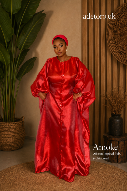 A woman modeling a vibrant red satin Amoke African-inspired dress with a matching red headwrap, standing in a styled indoor setting with a potted plant and wooden slats.