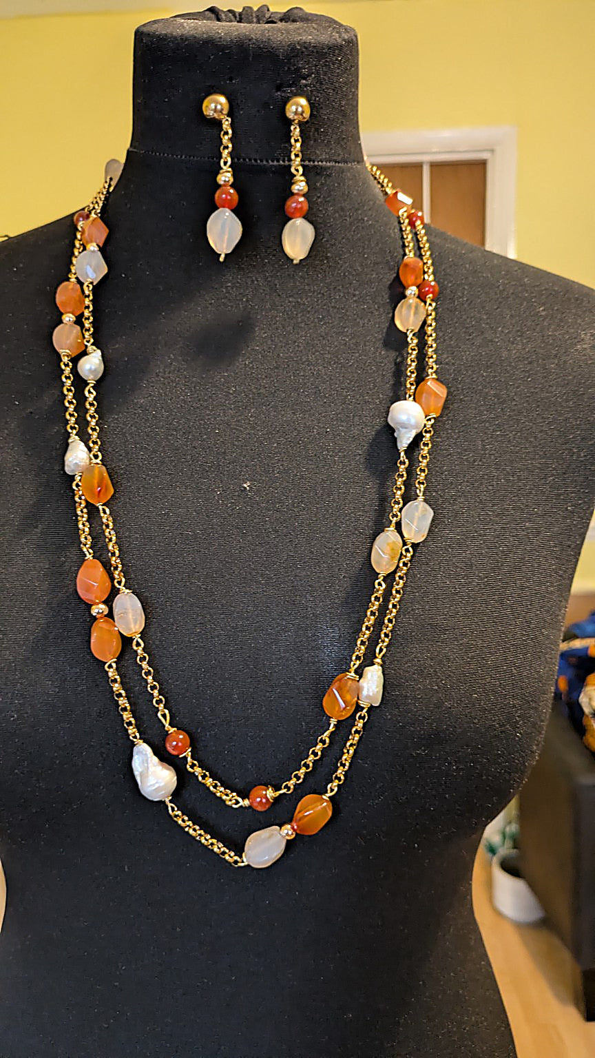 A double-strand gold chain necklace with orange and white agate beads, displayed on a black mannequin torso alongside matching drop earrings.