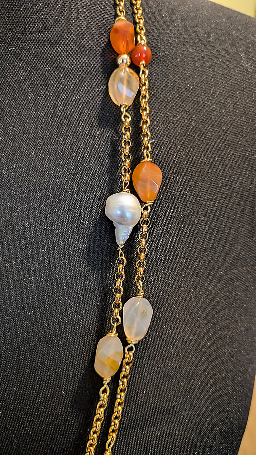 A close-up of a gold chain necklace displayed on a black mannequin bust, featuring amber-orange stone beads, a white baroque pearl, and translucent pale gray beads.