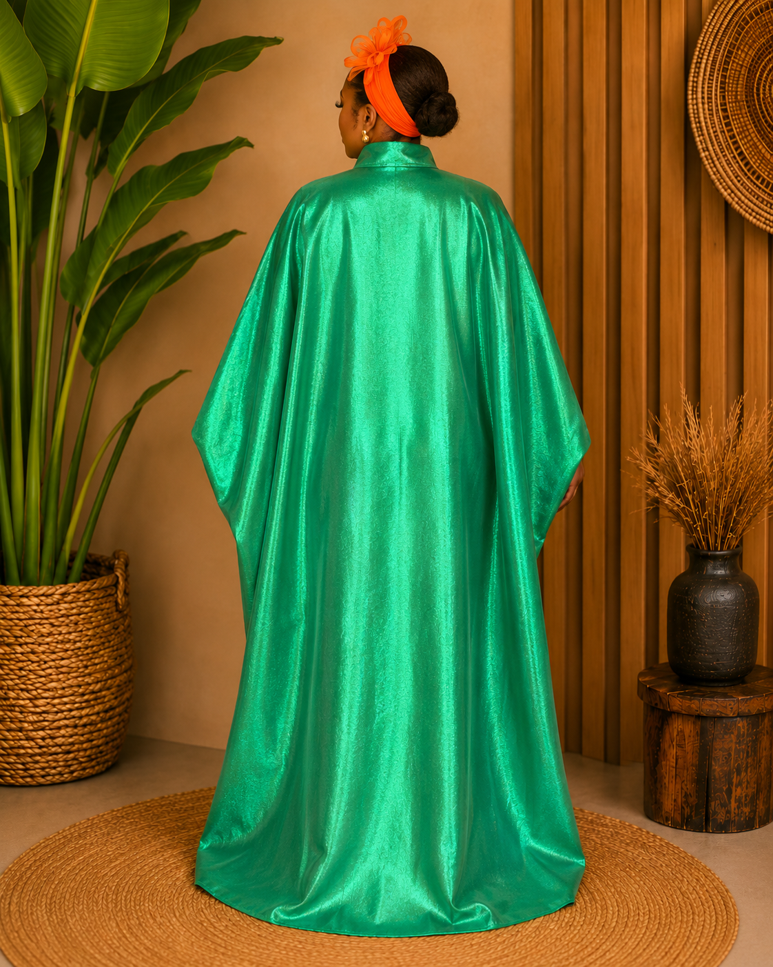 A woman viewed from behind wearing a flowing, floor-length emerald green satin or silk caftan with wide draped sleeves, styled in a warm-toned bohemian room with a potted plant and woven decor.