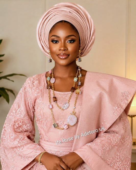 A woman wearing a light pink traditional outfit with matching headwrap, layered gold and stone necklaces, and earrings, posed against a softly lit indoor background.