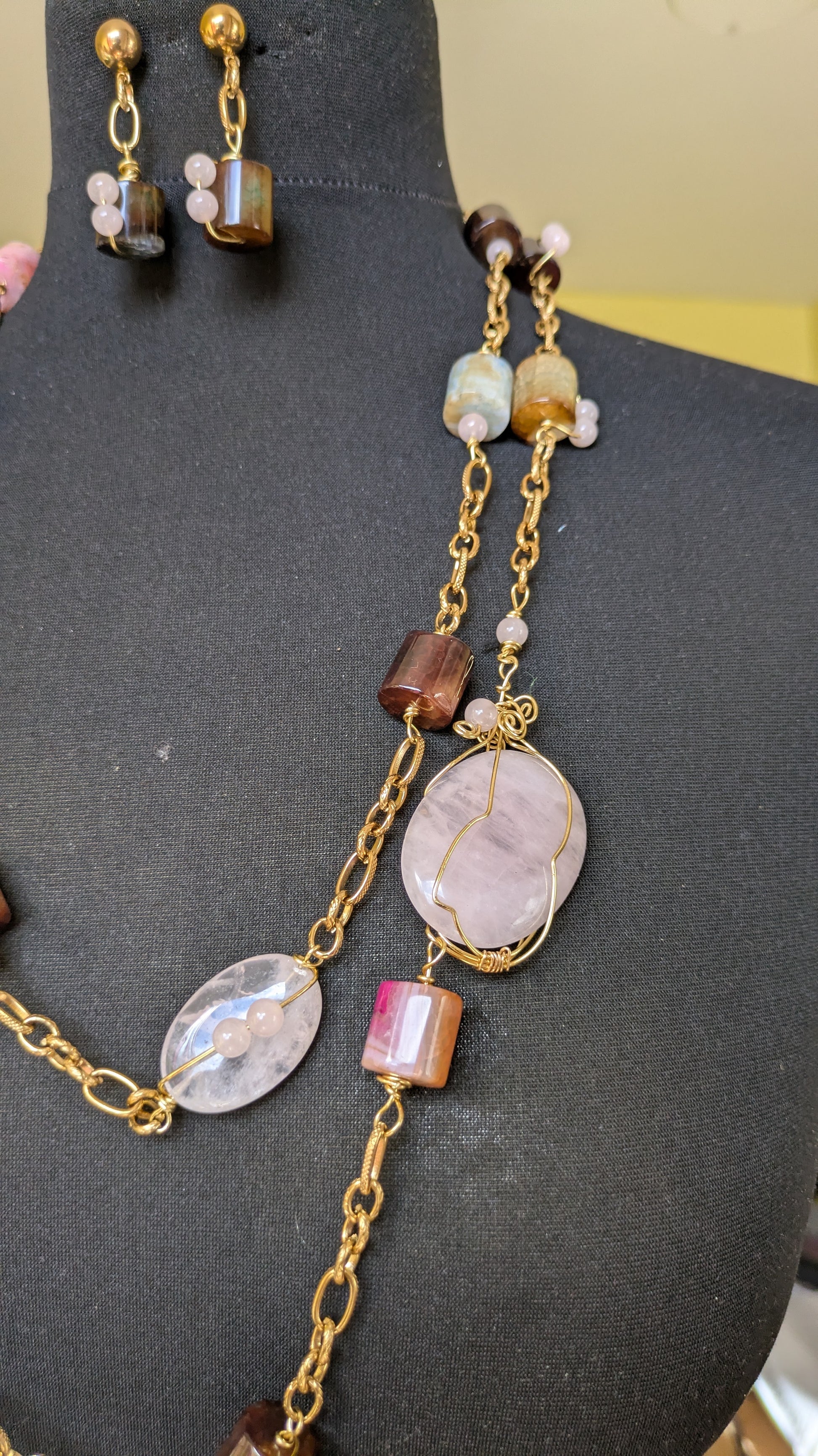 A gold-tone jewelry set displayed on a black mannequin bust, featuring a lariat-style necklace with a large oval pink stone pendant, smaller pink and brown beads, and matching drop earrings with gold studs and layered stones.